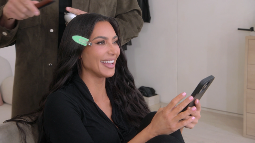 Kim Kardashian smiles as she holds her phone while fixing her hair.