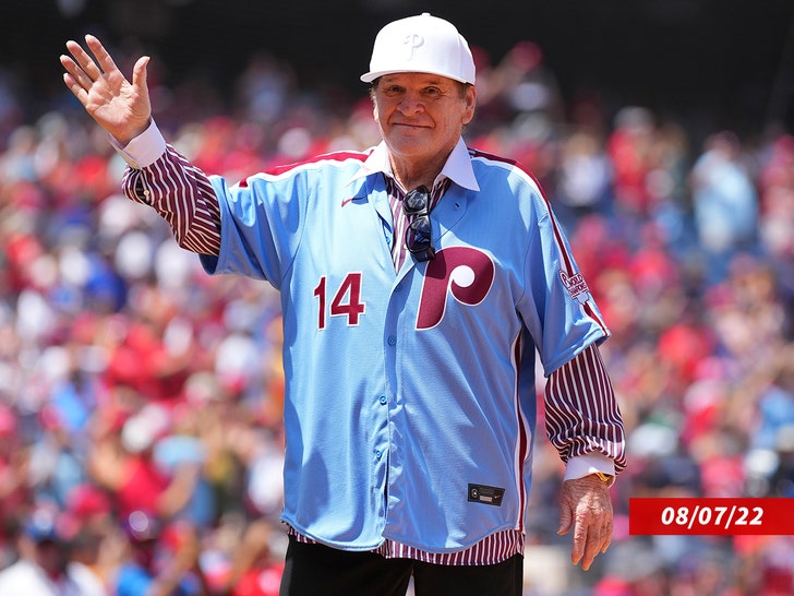 Pete Rose waving while wearing a striped shirt under a blue Philadelphia Phillies jersey and a white Phillies cap.